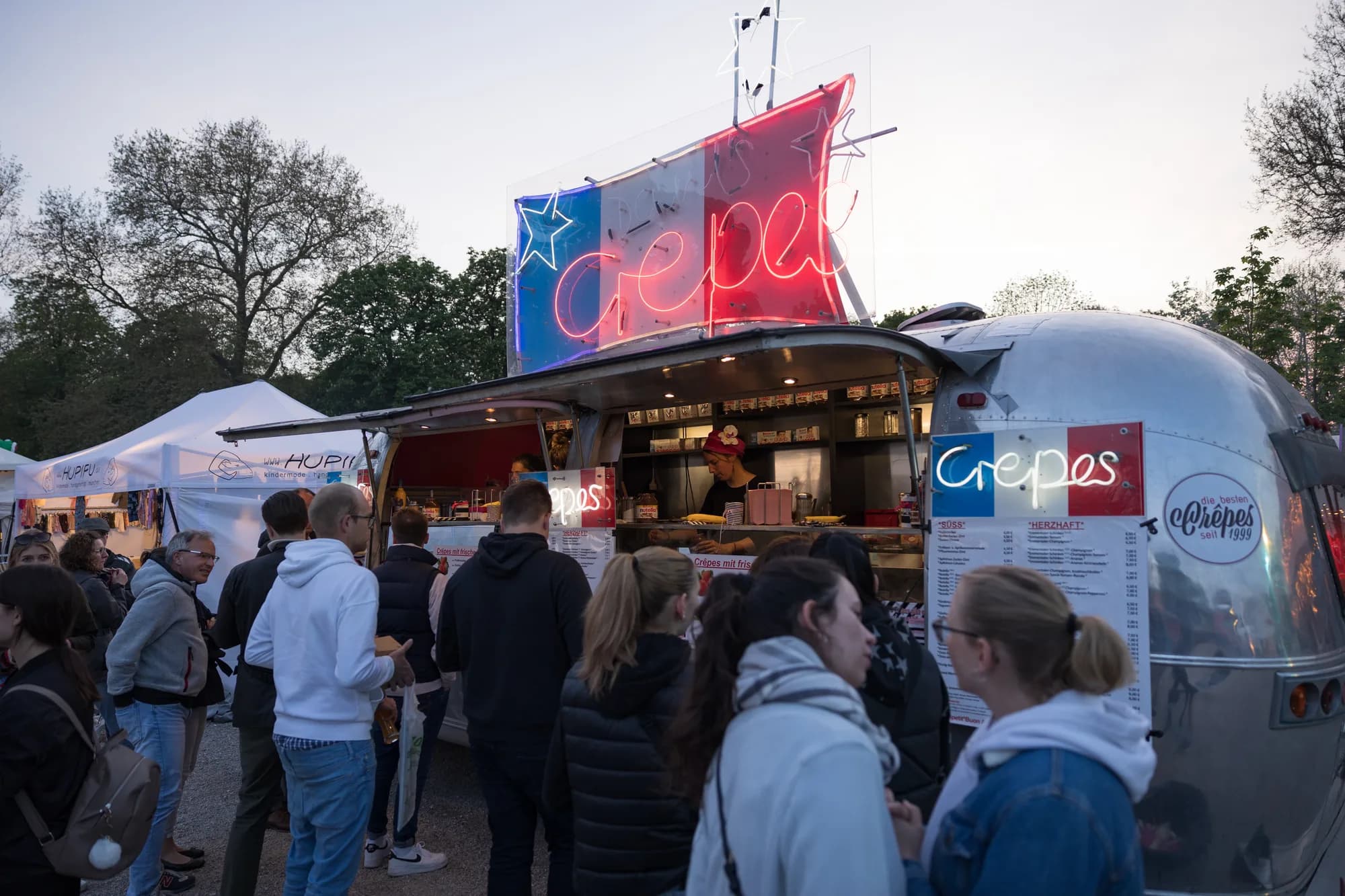 A silver Airstream trailer with a French flag neon sign serves crepes to a crowd of festival-goers at dusk.
