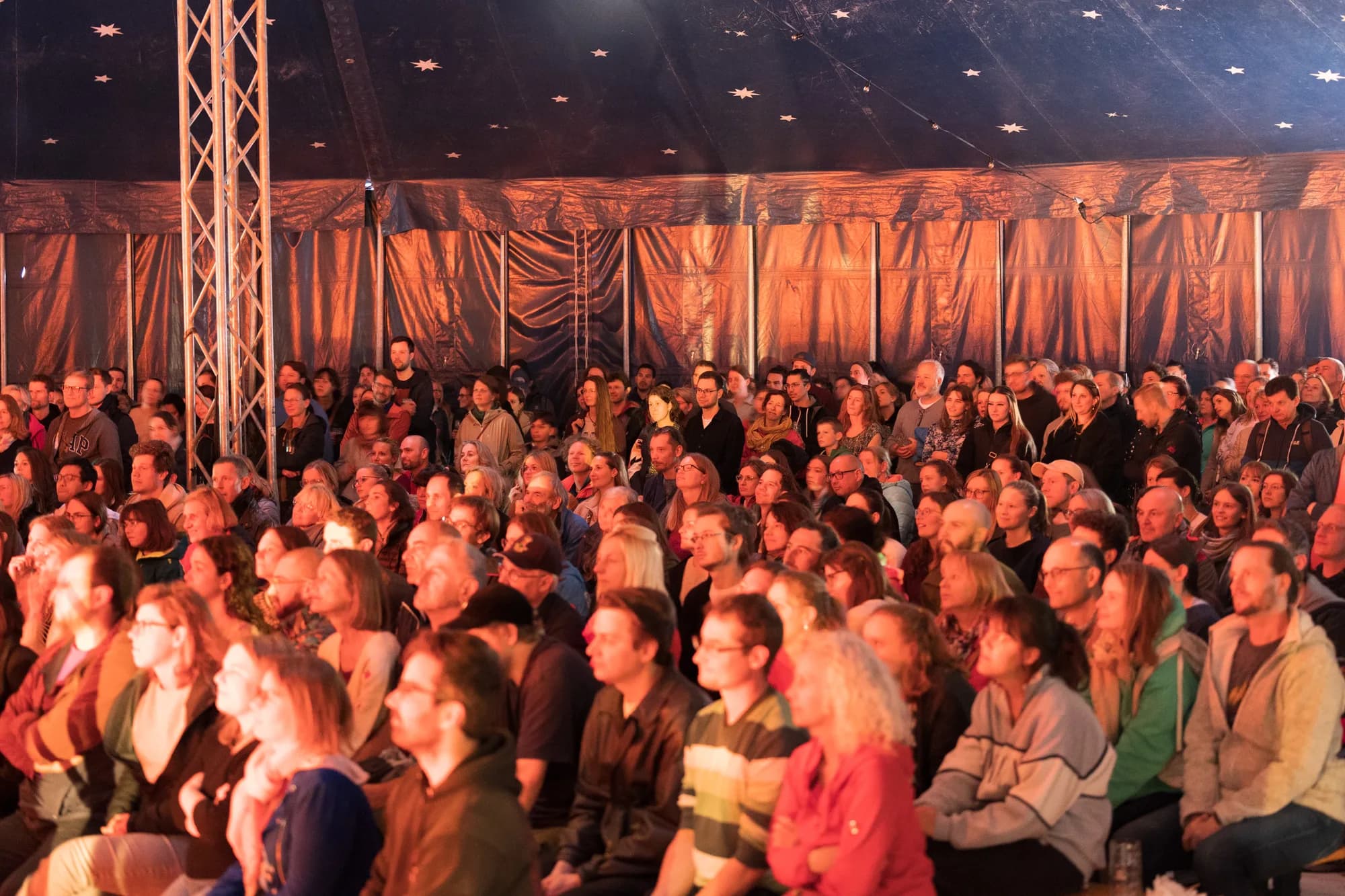 A large, diverse crowd of festival-goers sits closely together inside a tent, looking intently towards the left side of the frame.
