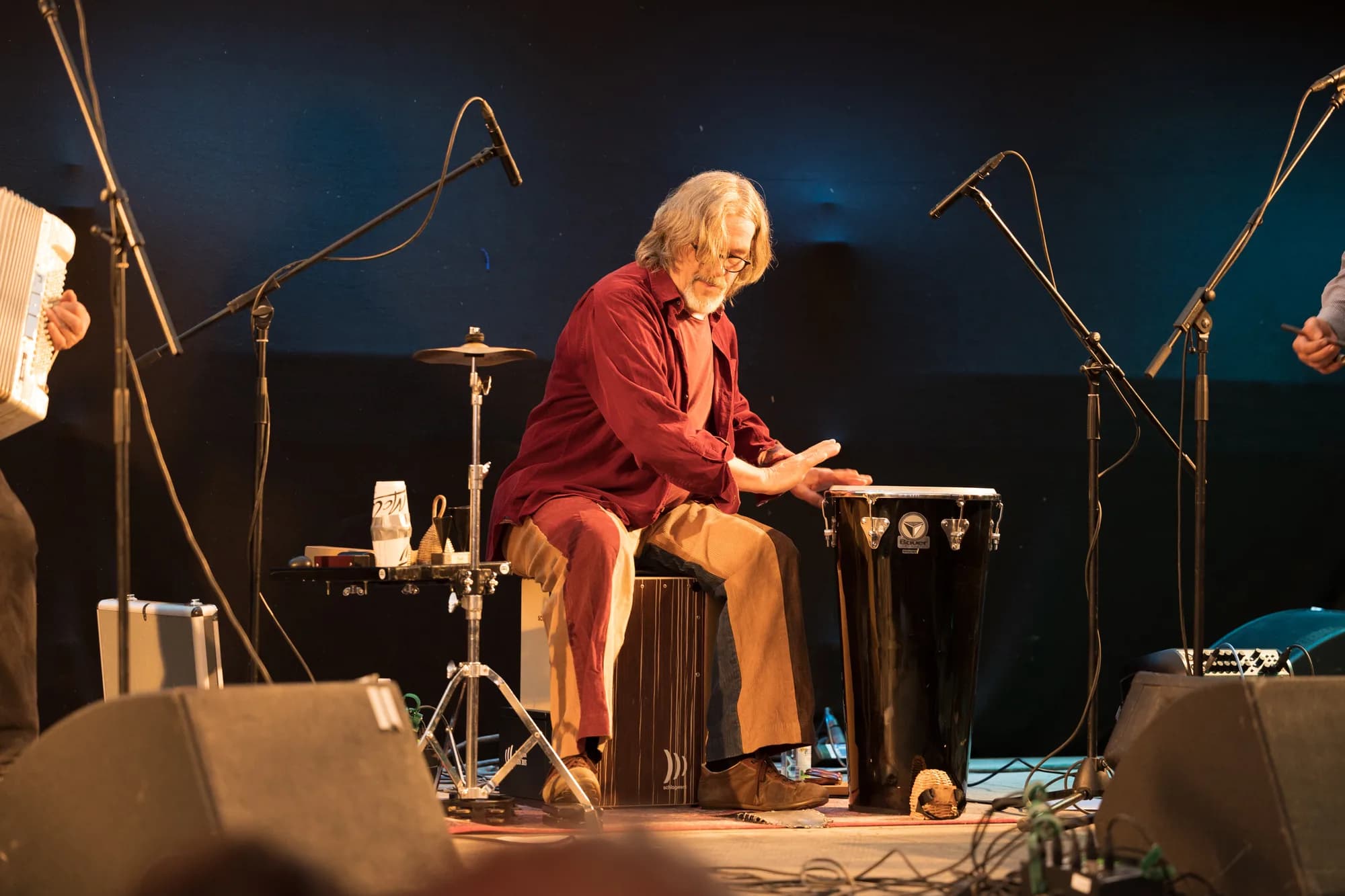 A musician with long grey hair and a red shirt plays a conga drum on a dimly lit stage.