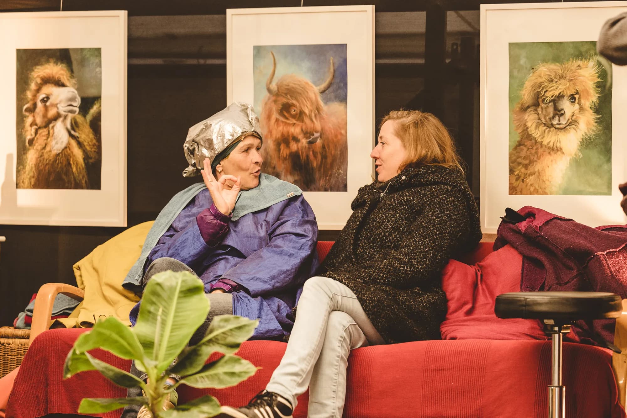 Two performers in eclectic costumes sit on a red sofa in front of three framed animal portraits.