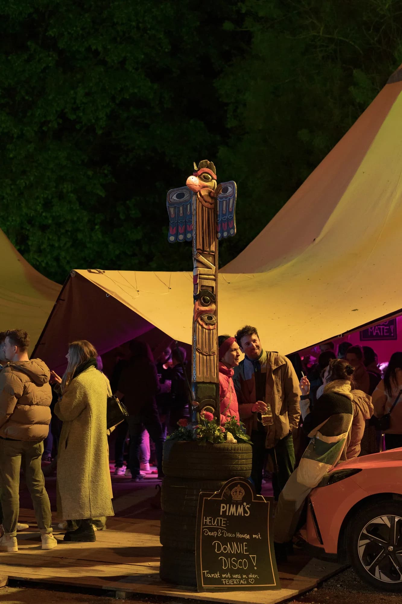 A wooden totem pole stands next to a chalkboard sign advertising Pimm's and Donnie Disco at a night market stall.