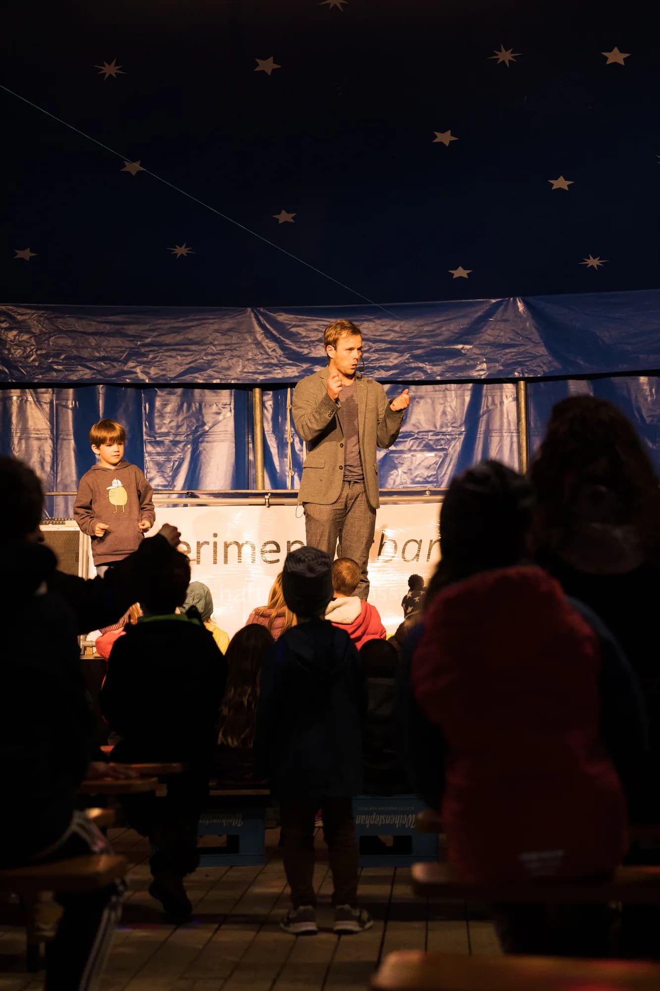 A performer in a grey suit and a young boy in a brown sweater stand on a wooden stage under a starry blue ceiling, entertaining a seated audience of children.