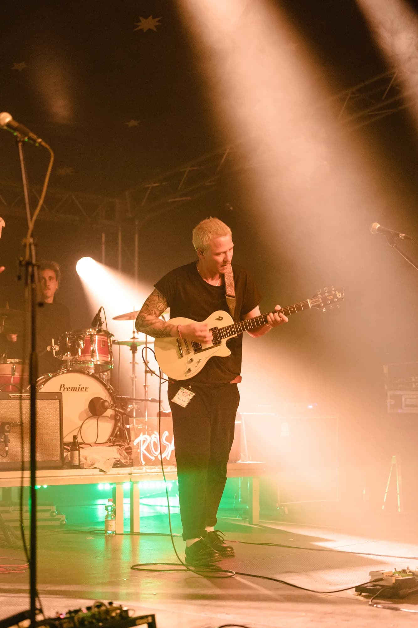 A guitarist with blonde hair and tattoos plays a white electric guitar on a stage illuminated by bright spotlights and haze.