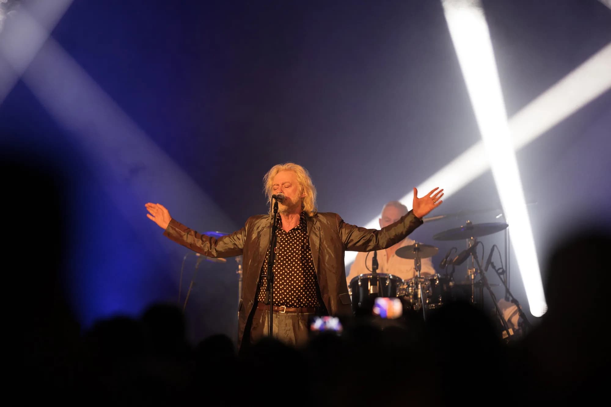 A male singer with blonde hair and a leather jacket performs on stage with arms wide open under bright spotlights.