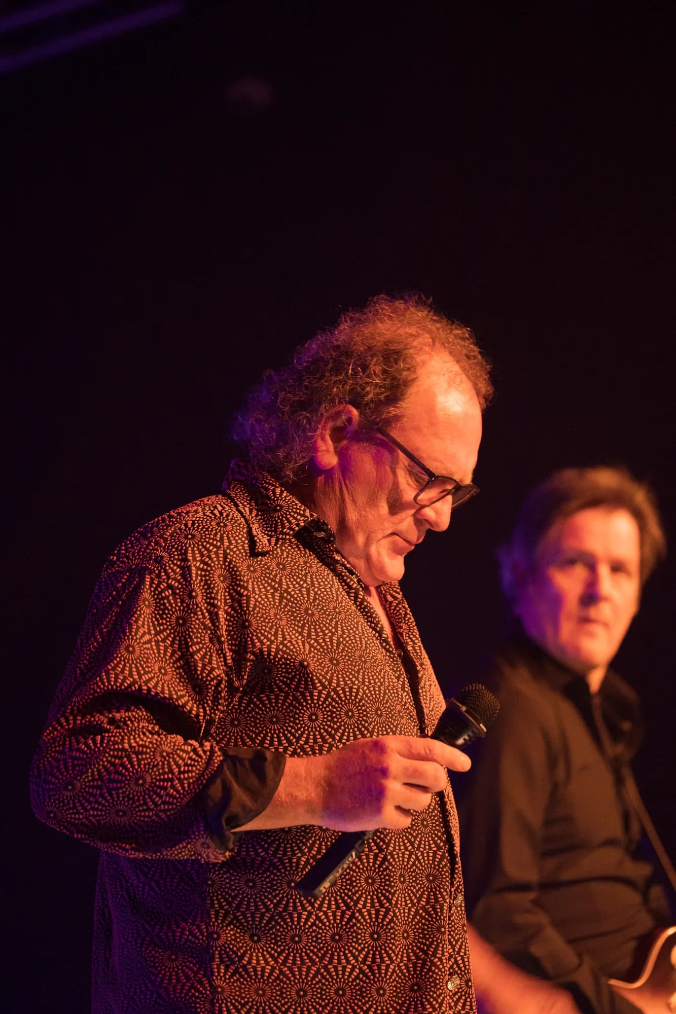 A male singer in a patterned shirt speaks into a microphone on a dimly lit stage while a bandmate sits in the background.