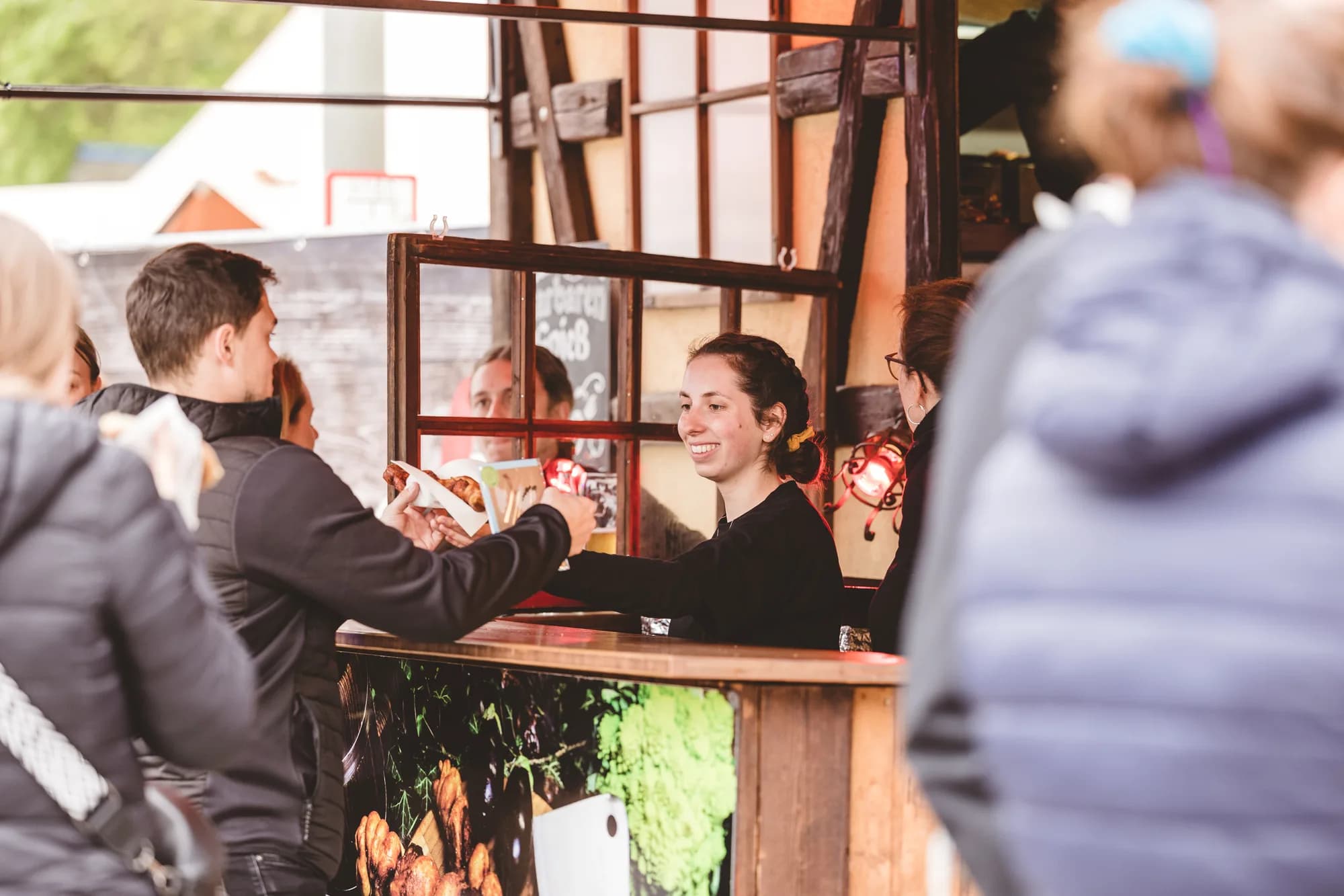 A smiling female server hands a wrapped food item to a customer at a wooden festival stall.