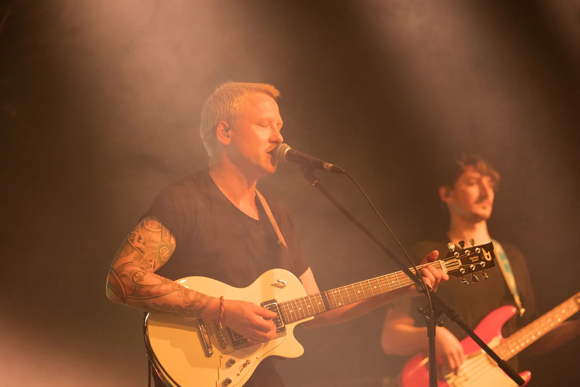 A male musician with a tattooed arm plays a white electric guitar and sings into a microphone on a dimly lit stage.
