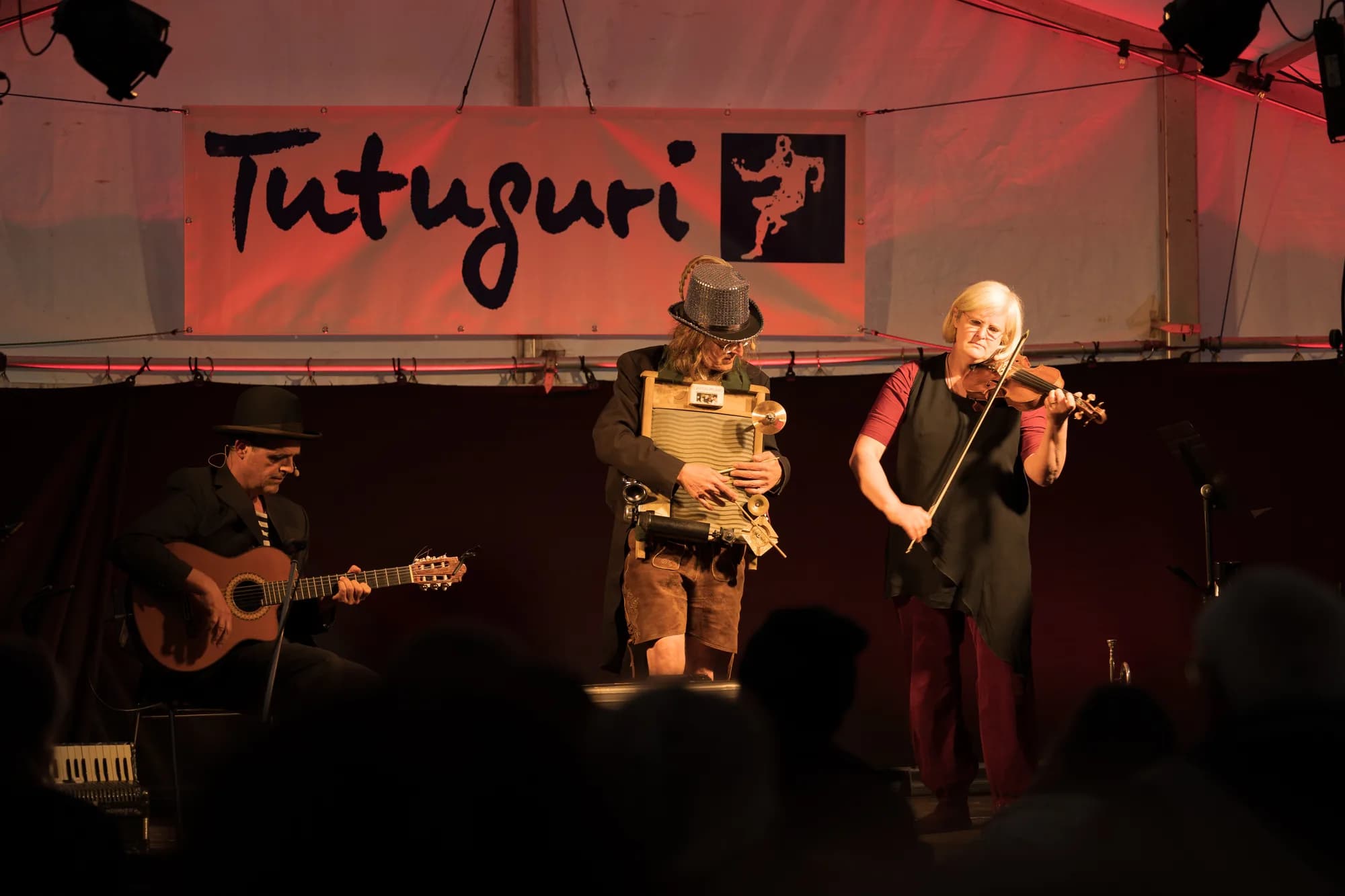 A folk trio performs on stage with a guitarist, accordion player, and singer under a red banner.