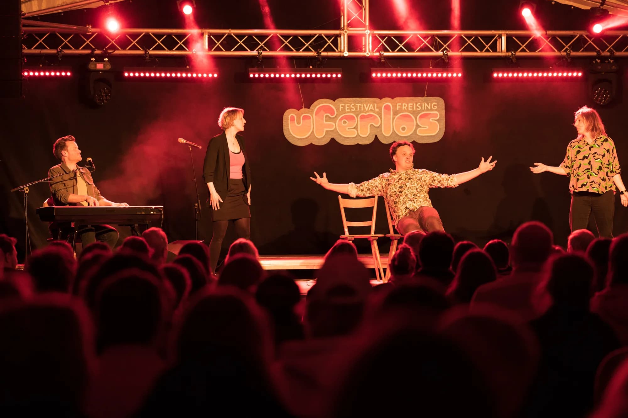 A four-person comedy troupe performs on stage under red lights with the Uferlos Freising logo behind them.