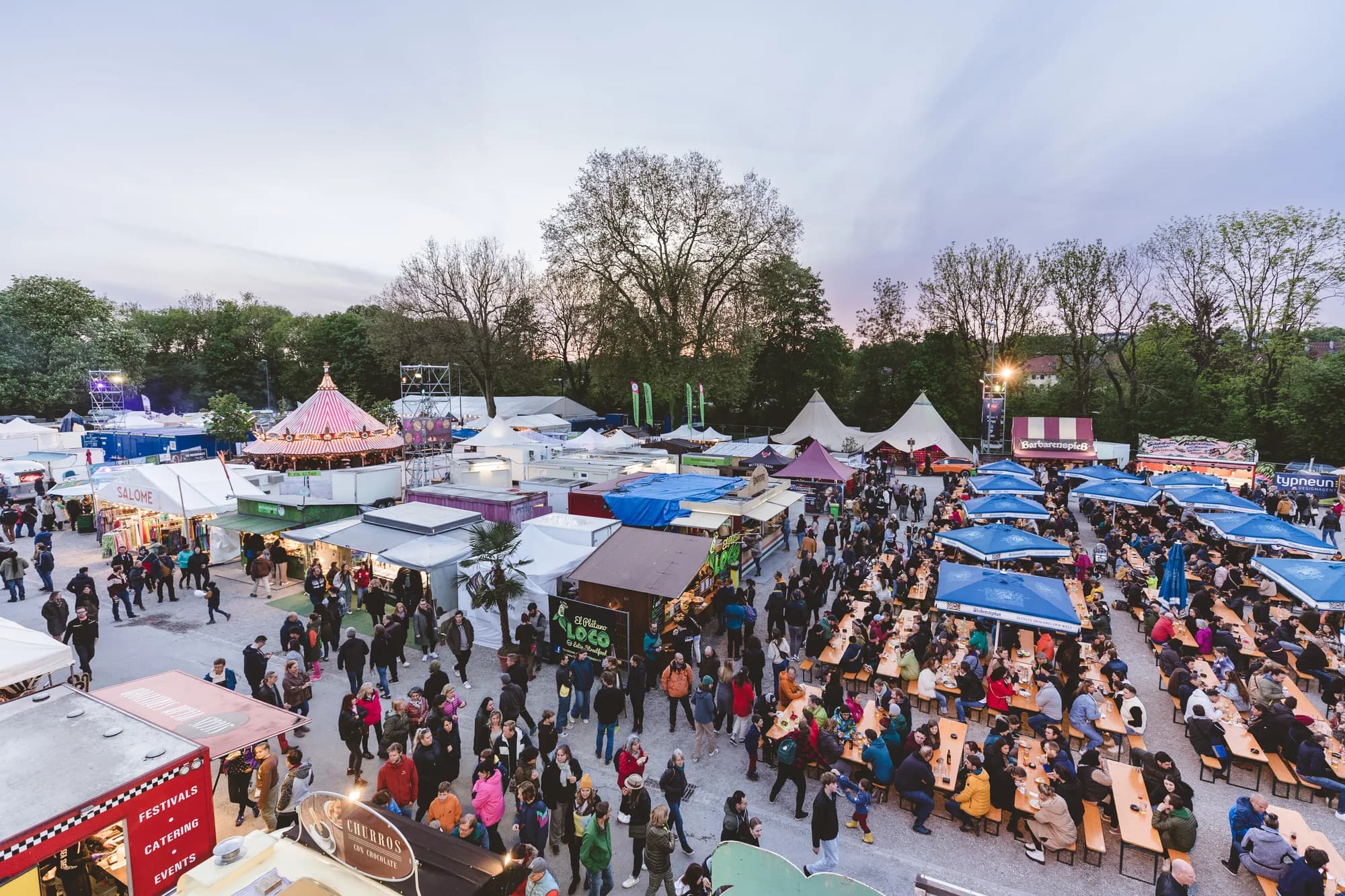 A high-angle drone shot of the Uferlos festival grounds featuring large red circus tents and a crowded market area with the Freising cathedral in the background.