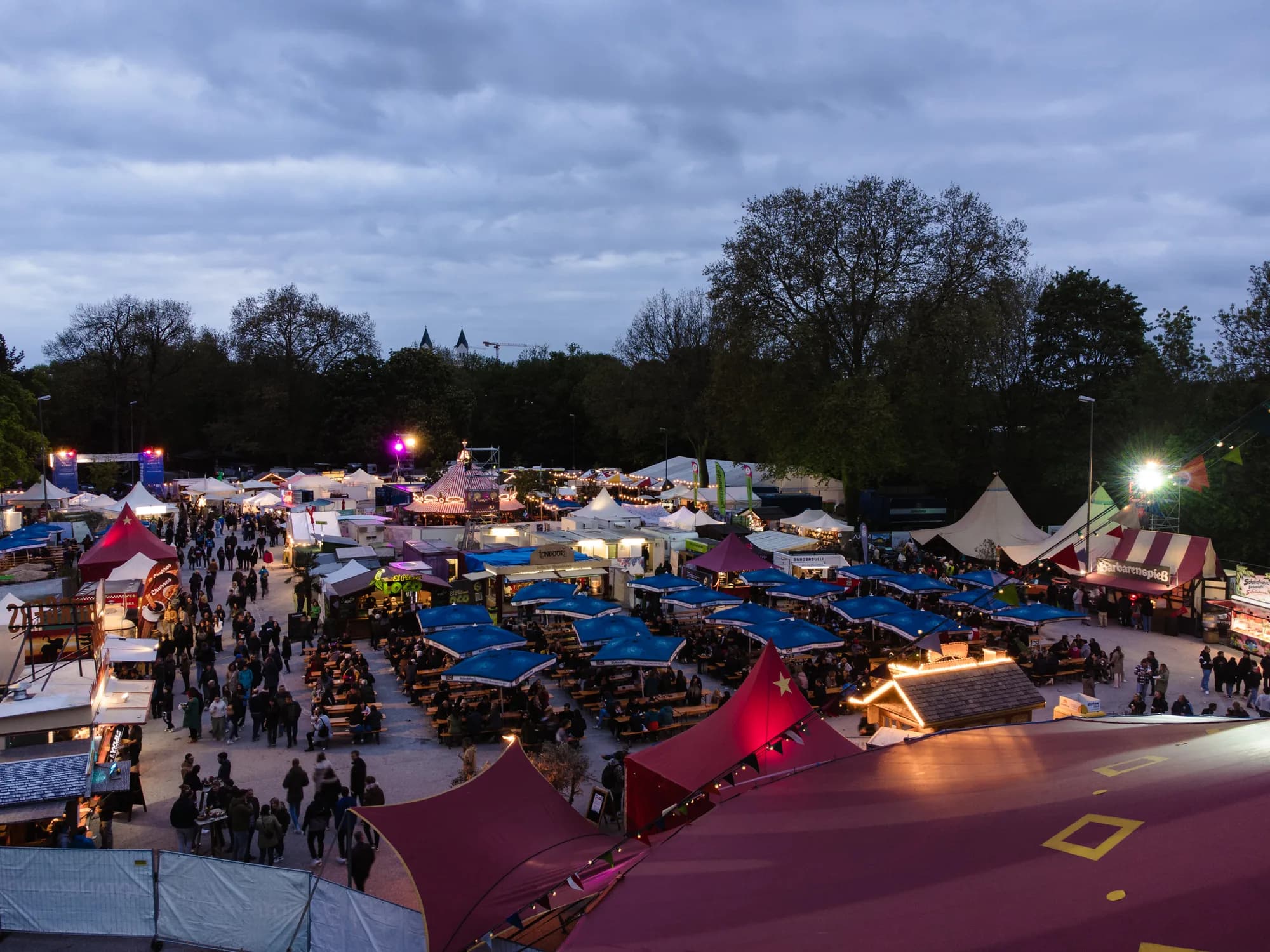 A high-angle drone shot of the Uferlos festival grounds at dusk, showing crowded market stalls and tents under a cloudy sky.