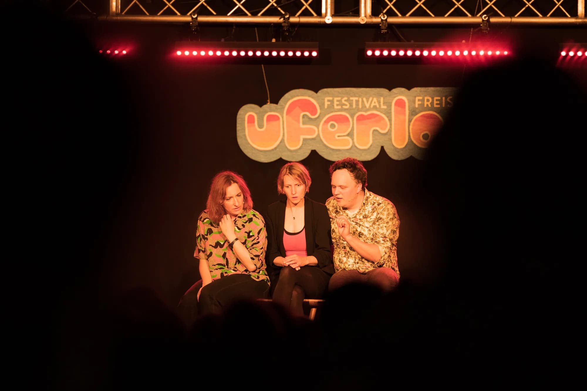 Three speakers seated on a stage in front of a Uferlos banner, framed by the silhouettes of an attentive audience.