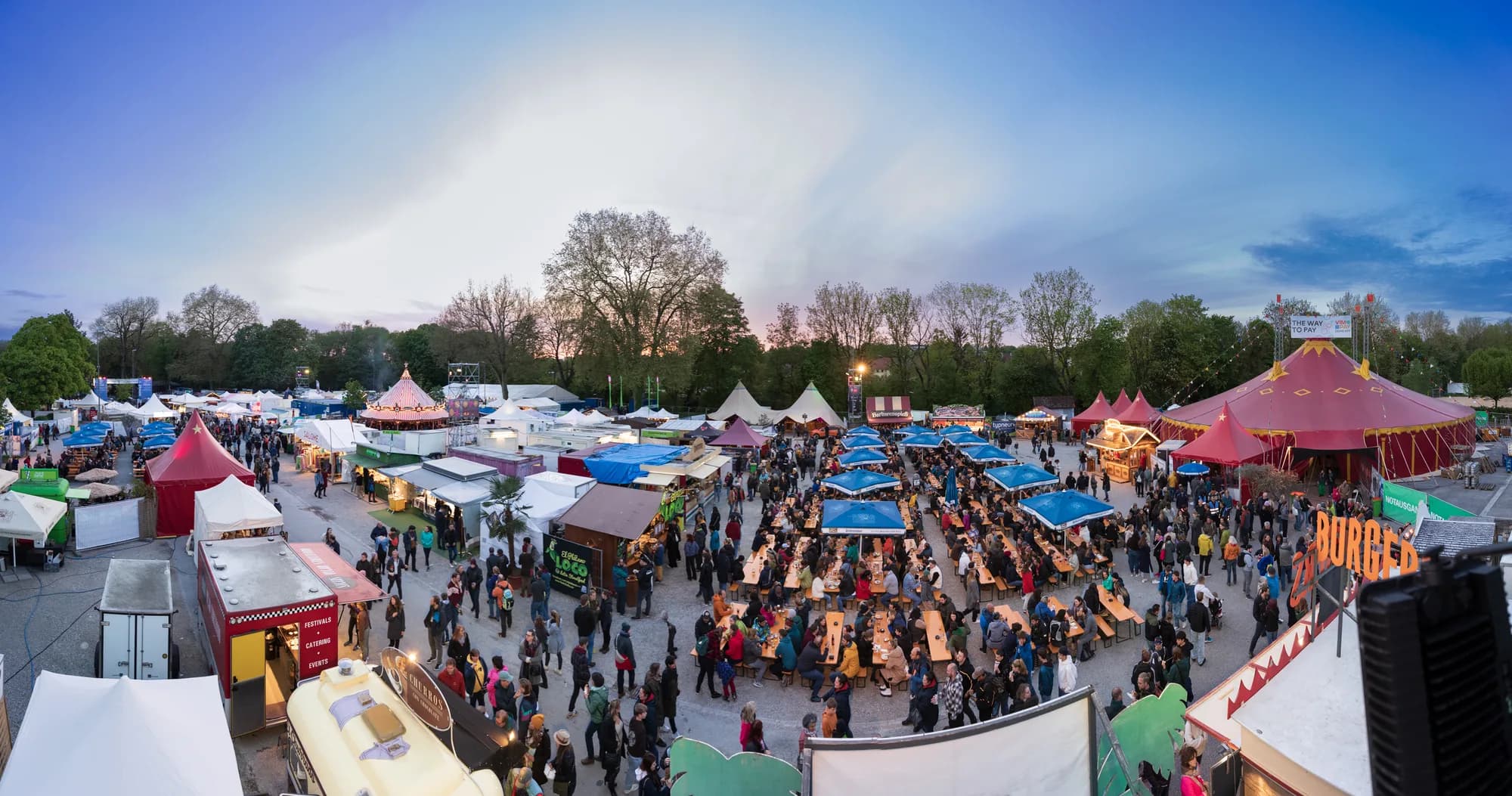 A high-angle view of the crowded festival grounds at dusk with colorful tents and warm light.