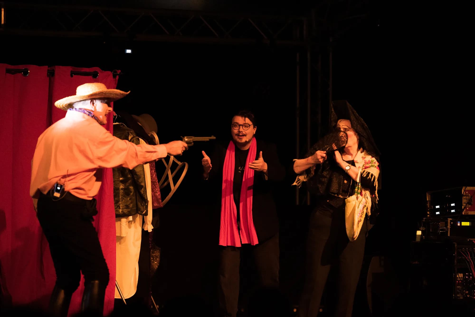 Three performers in western costumes act out a scene on stage with a red curtain backdrop.