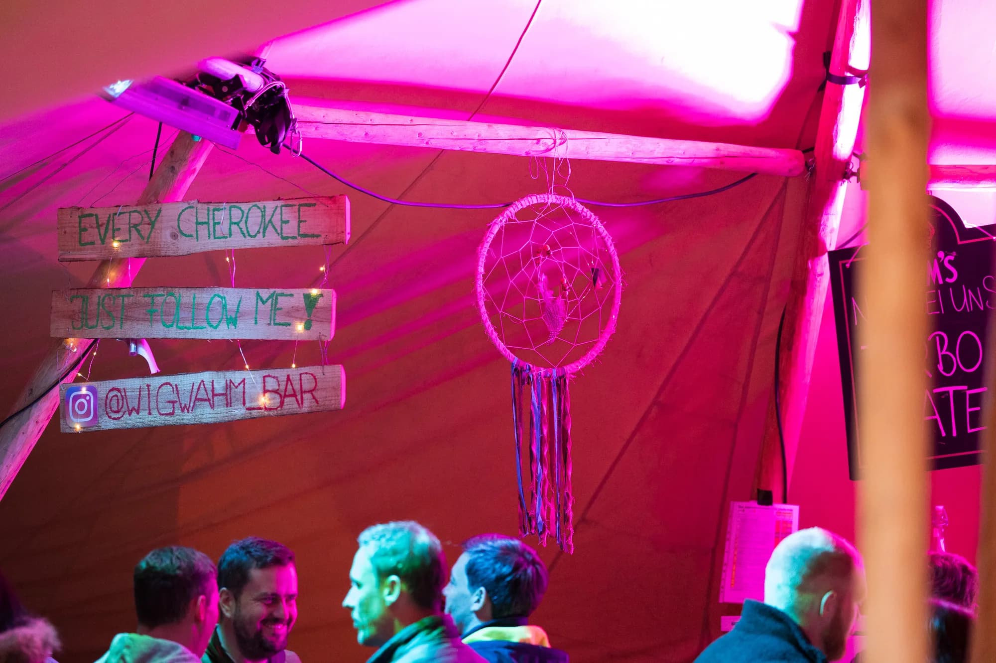 Hand-painted wooden signs inside the Wigwam Bar tent illuminated by warm pink light.