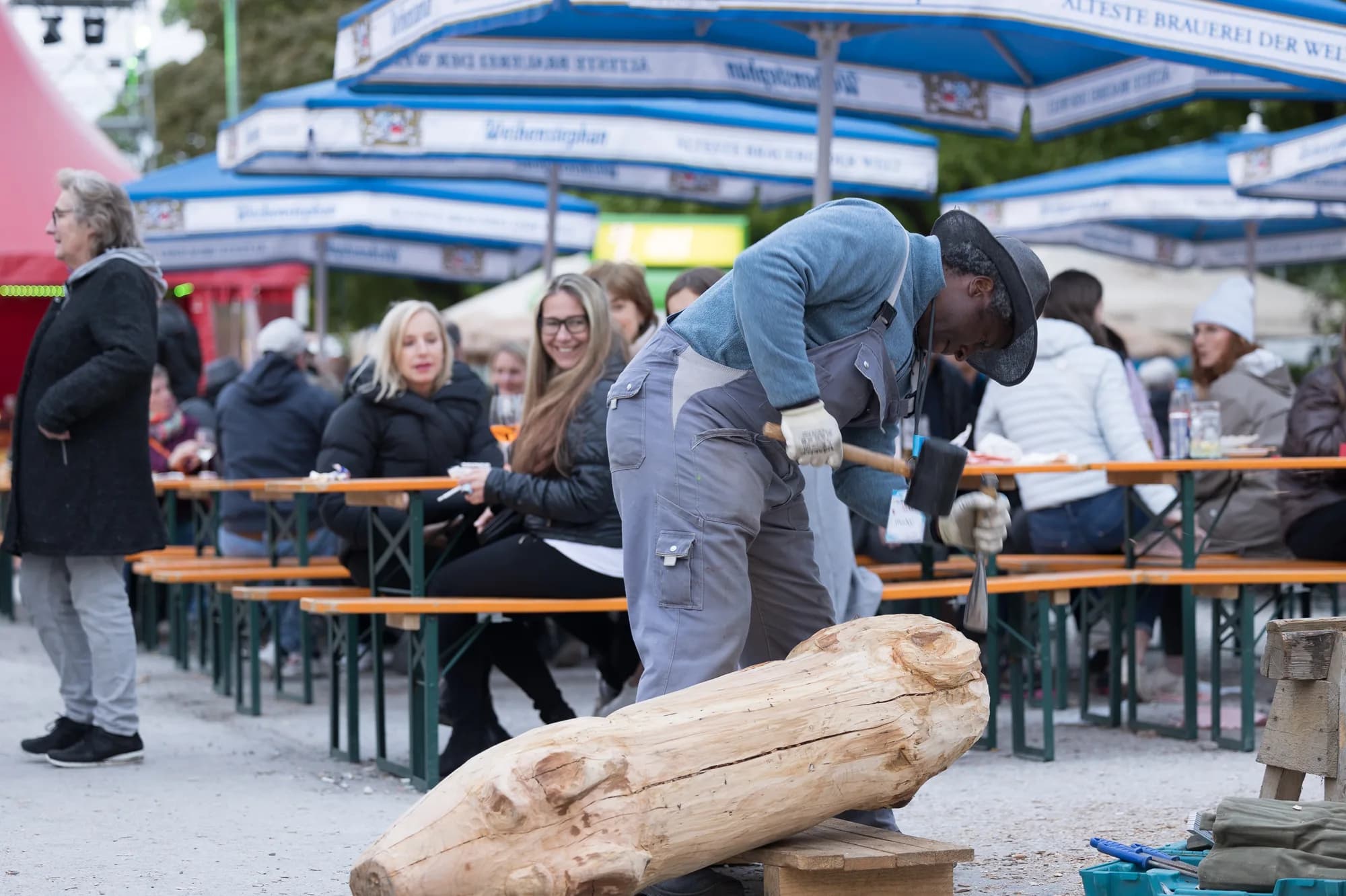 A woodcarver in a blue shirt and hat uses a mallet and chisel to shape a log in front of seated festival guests.
