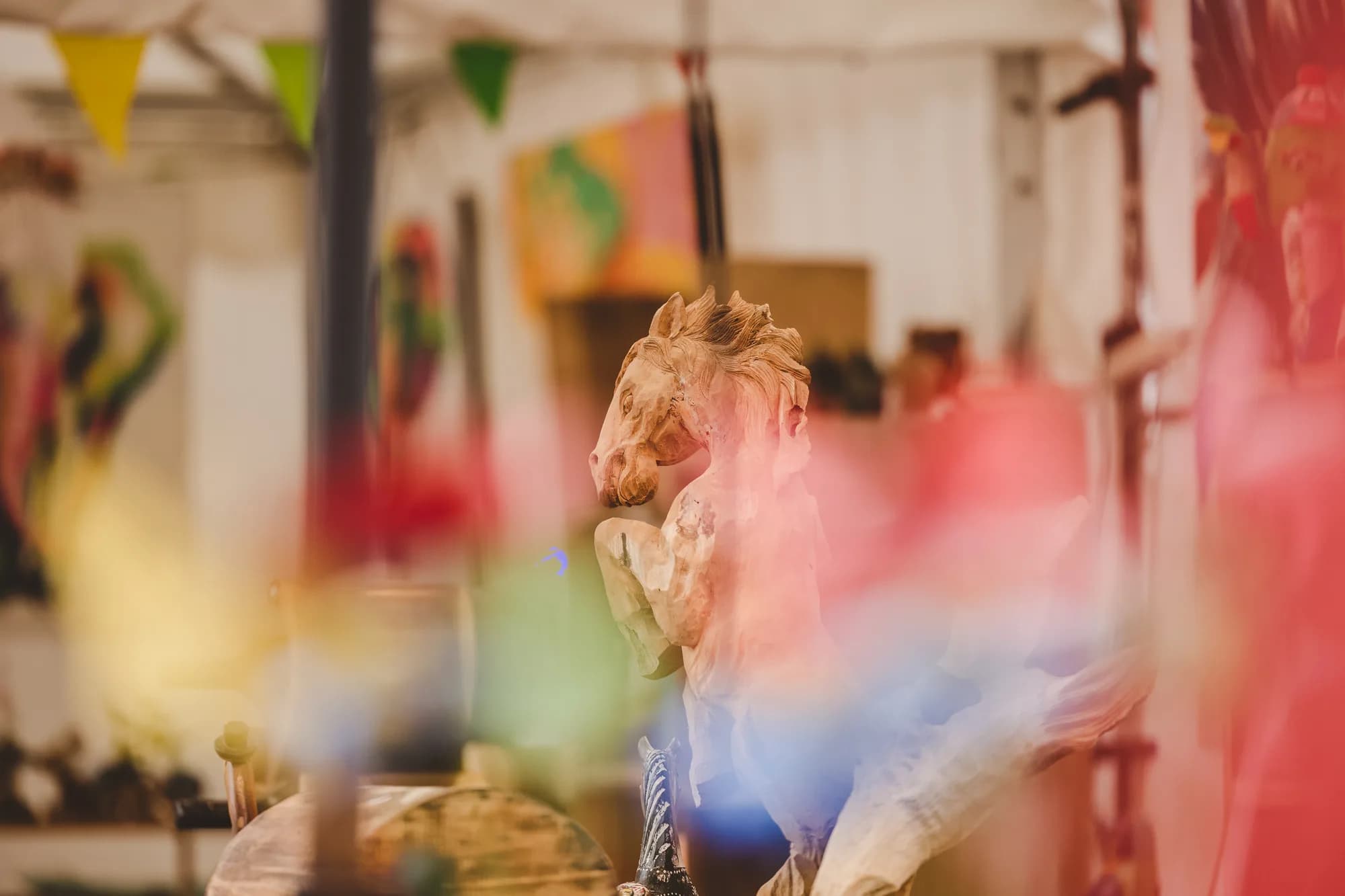 A wooden carved horse figurine stands on a market stall table with colorful blurred items in the foreground.