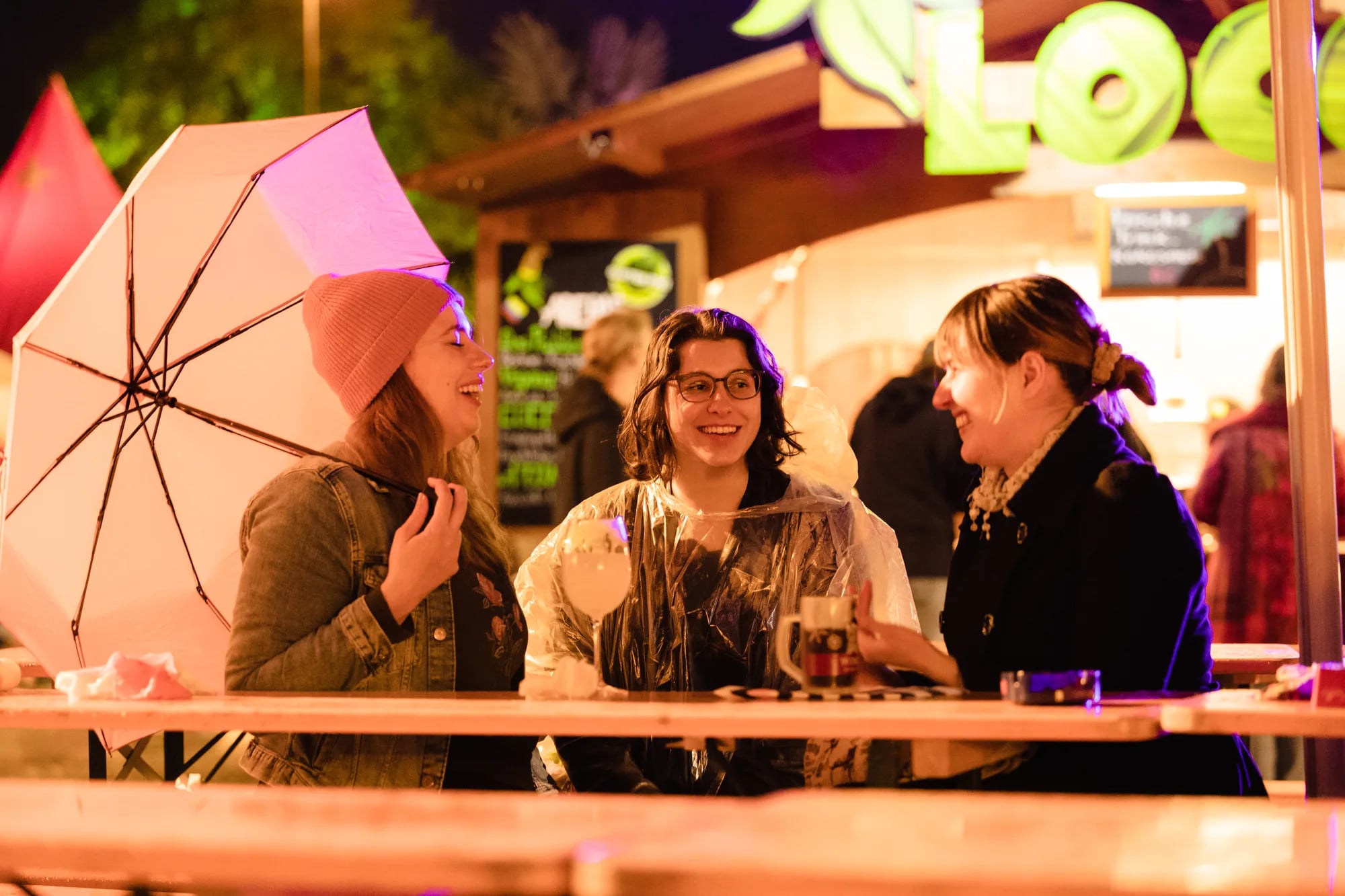 Drei lachende Frauen sitzen bei Regen an einer Holzbar, eine hält einen Regenschirm